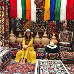 Woman in a yellow dress sitting among colorful rugs in a traditional carpet market